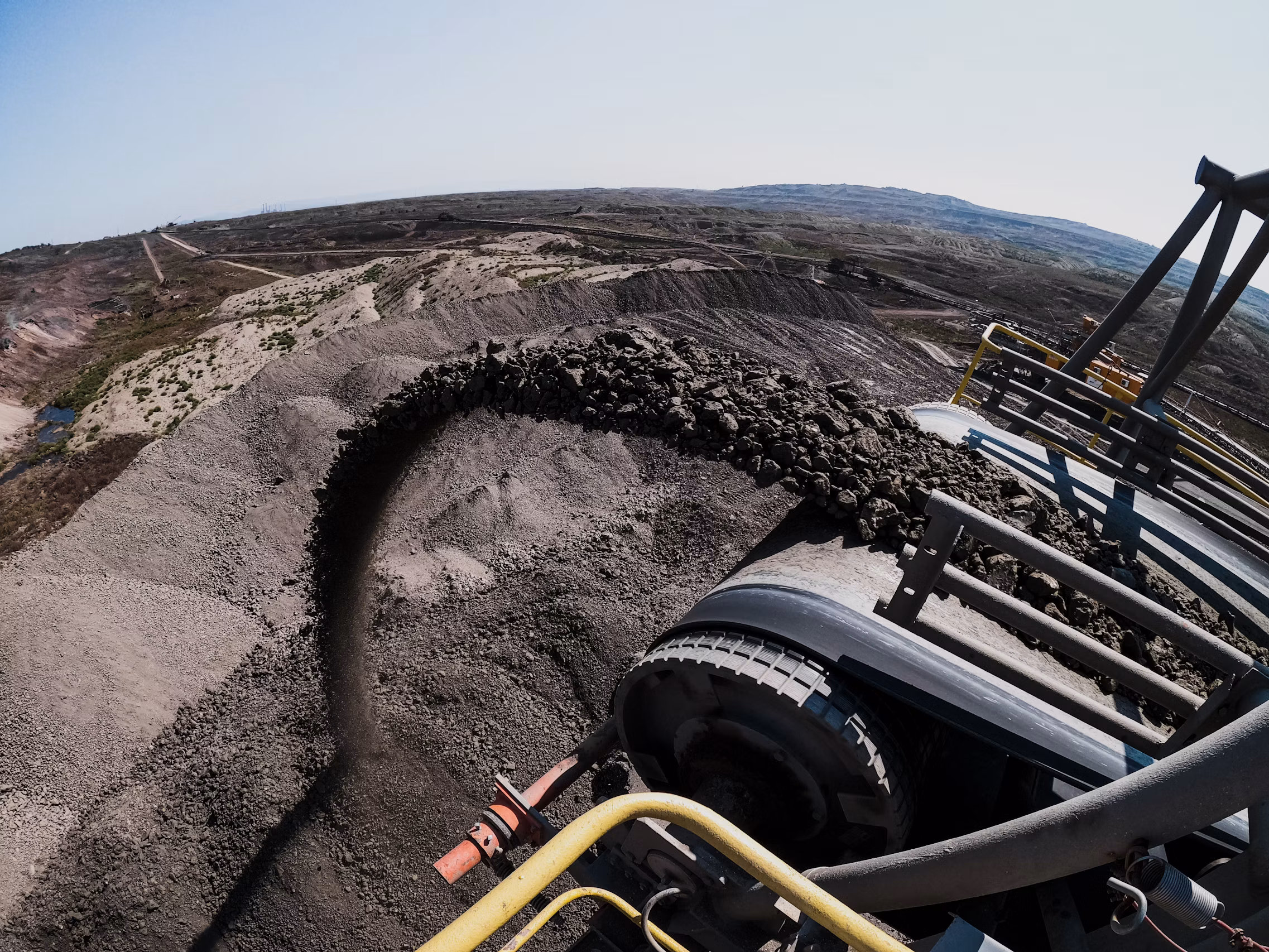 High-speed conveyor discharge pulley streaming mineral lumps onto terminal stockpile with open pit view